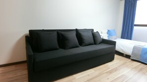 Ein schwarzes Sofa mit vier schwarzen Kissen in einem Raum mit Holzboden und einem Bett mit blau-weißer Bettdecke.