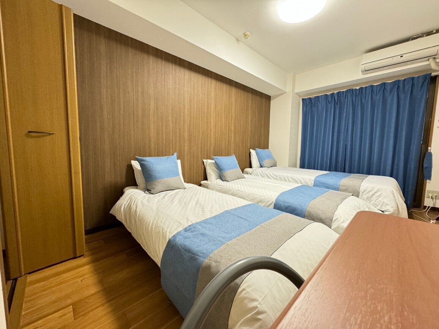 A hotel room with two beds, a wooden wardrobe, a desk, and a window with curtains.