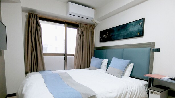 Ein Hotelzimmer mit Bett, Nachttisch, Klimaanlage, Fenster mit Vorhängen und einem Gemälde an der Wand.