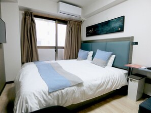 A hotel room with a bed, a nightstand, an air conditioning unit, a window with curtains, and a painting on the wall.