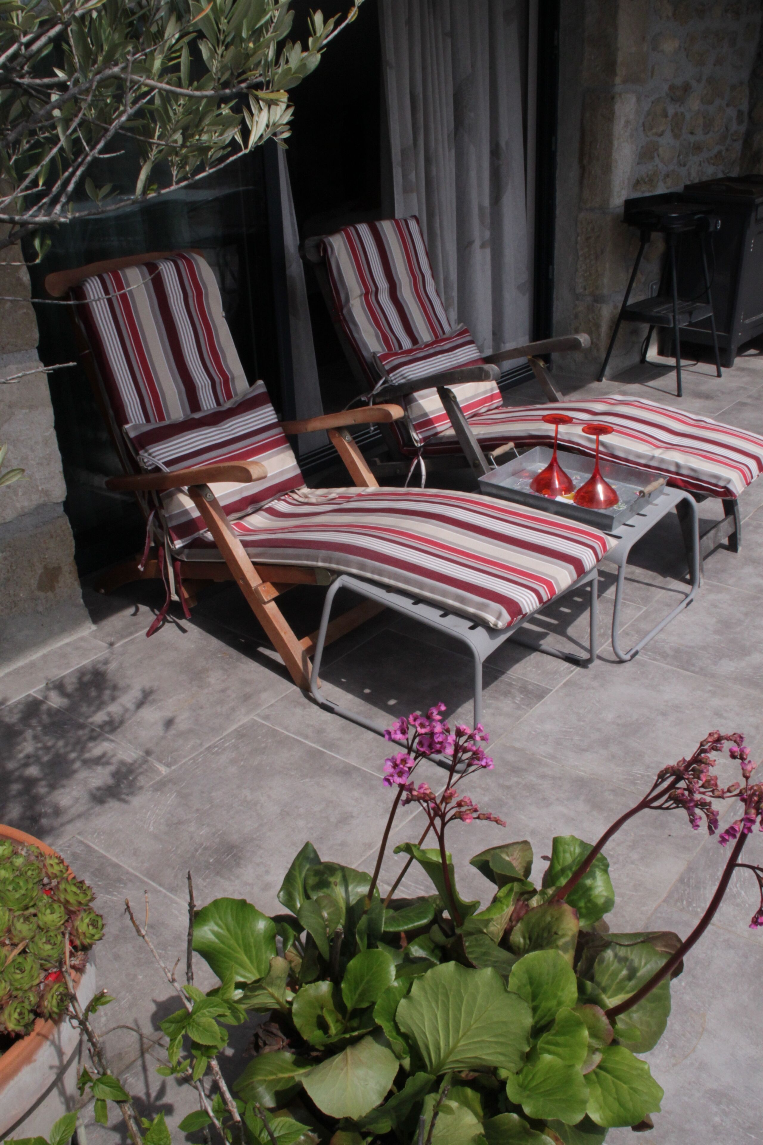 A patio area with two striped lounge chairs and a small table, surrounded by potted plants and a stone wall.