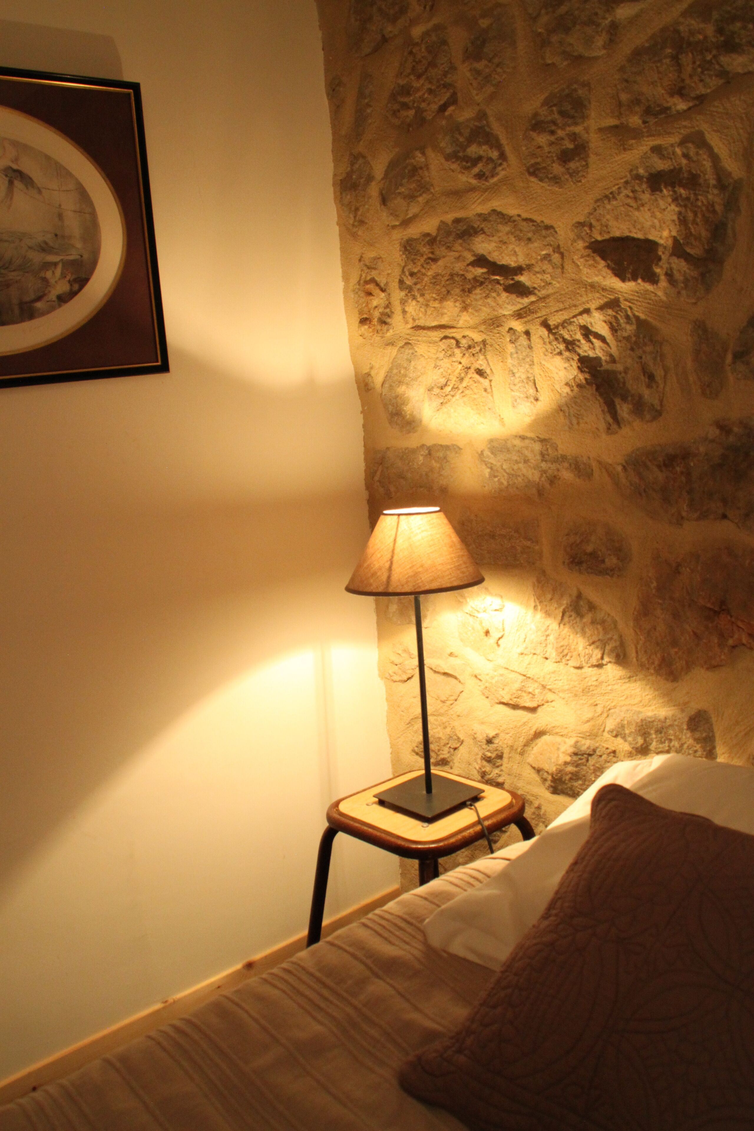 Un dormitorio con pared de piedra, una lámpara de noche, un cuadro enmarco y una cama con almohada.
