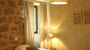 Una habitación con dos camas, una pared de piedra, una ventana con cortinas, una lámpara y cuadros enmarcados en la pared.