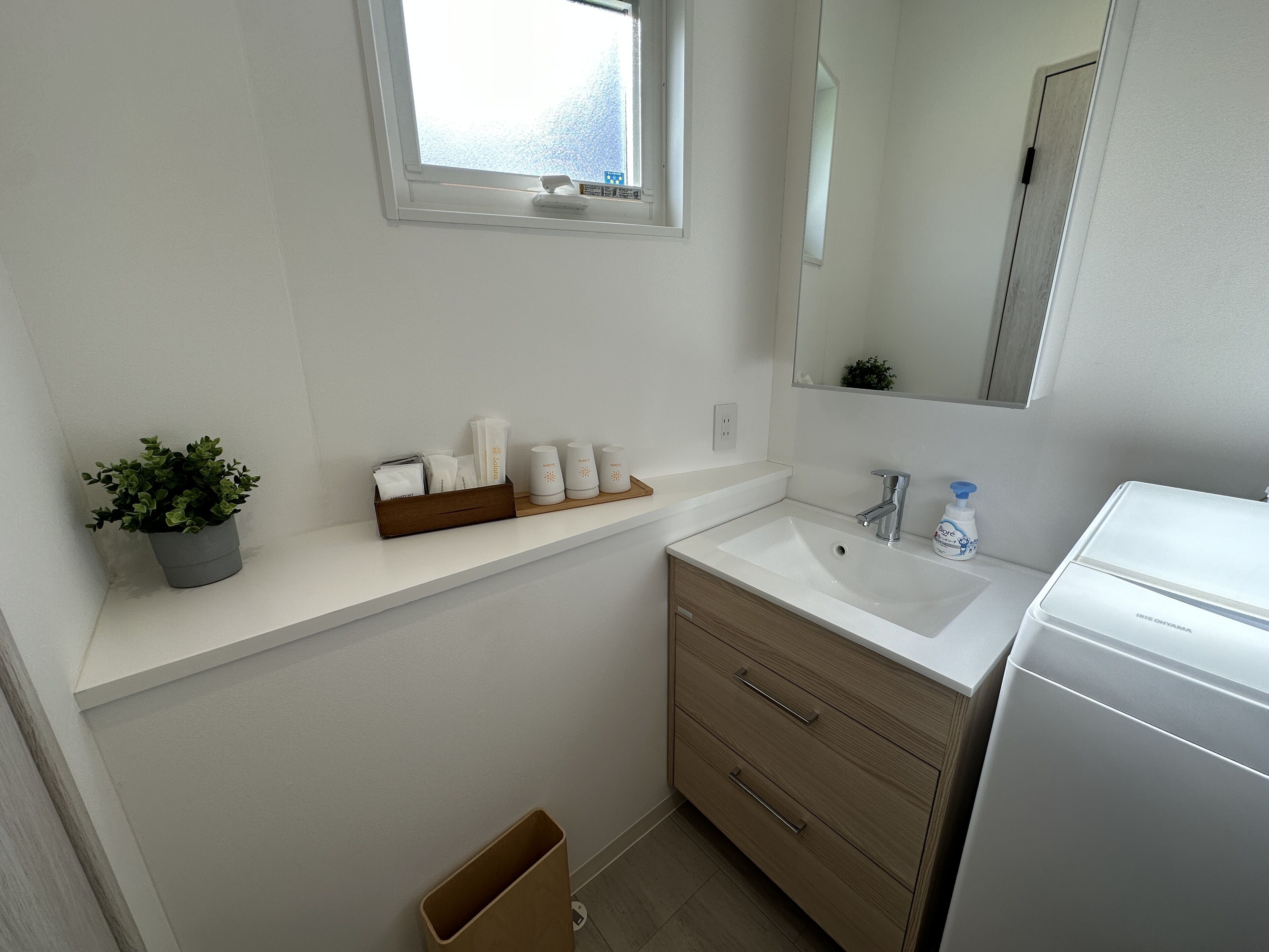 A bathroom with a white sink, wooden vanity, a mirror, a potted plant, and toiletries on the countertop.