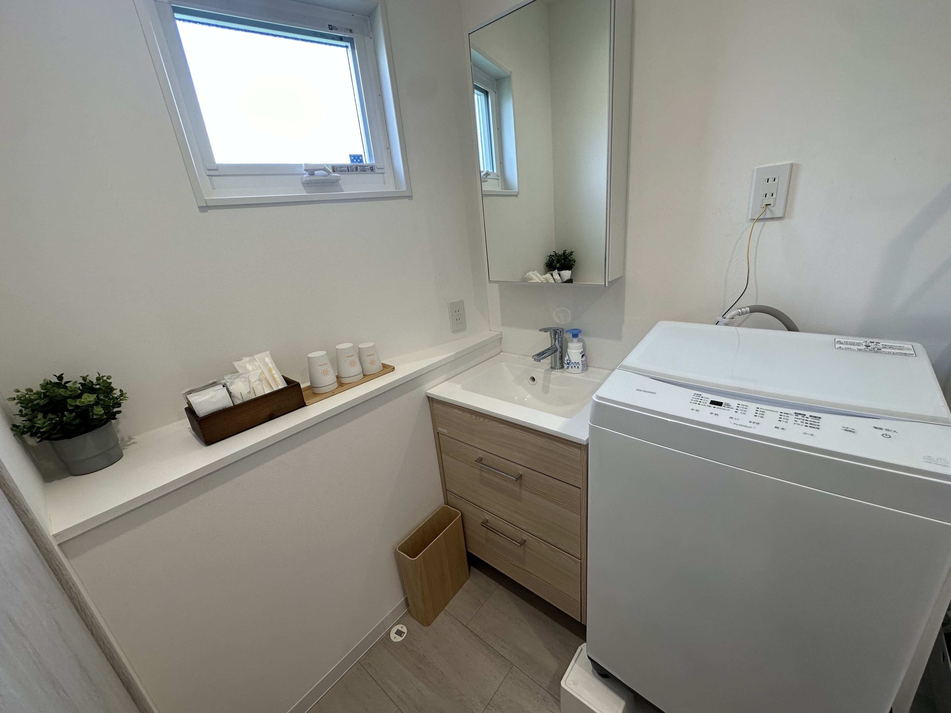 Un baño con un lavabo blanco, un espejo, una ventana, una planta pequeña y una lavadora blanca.