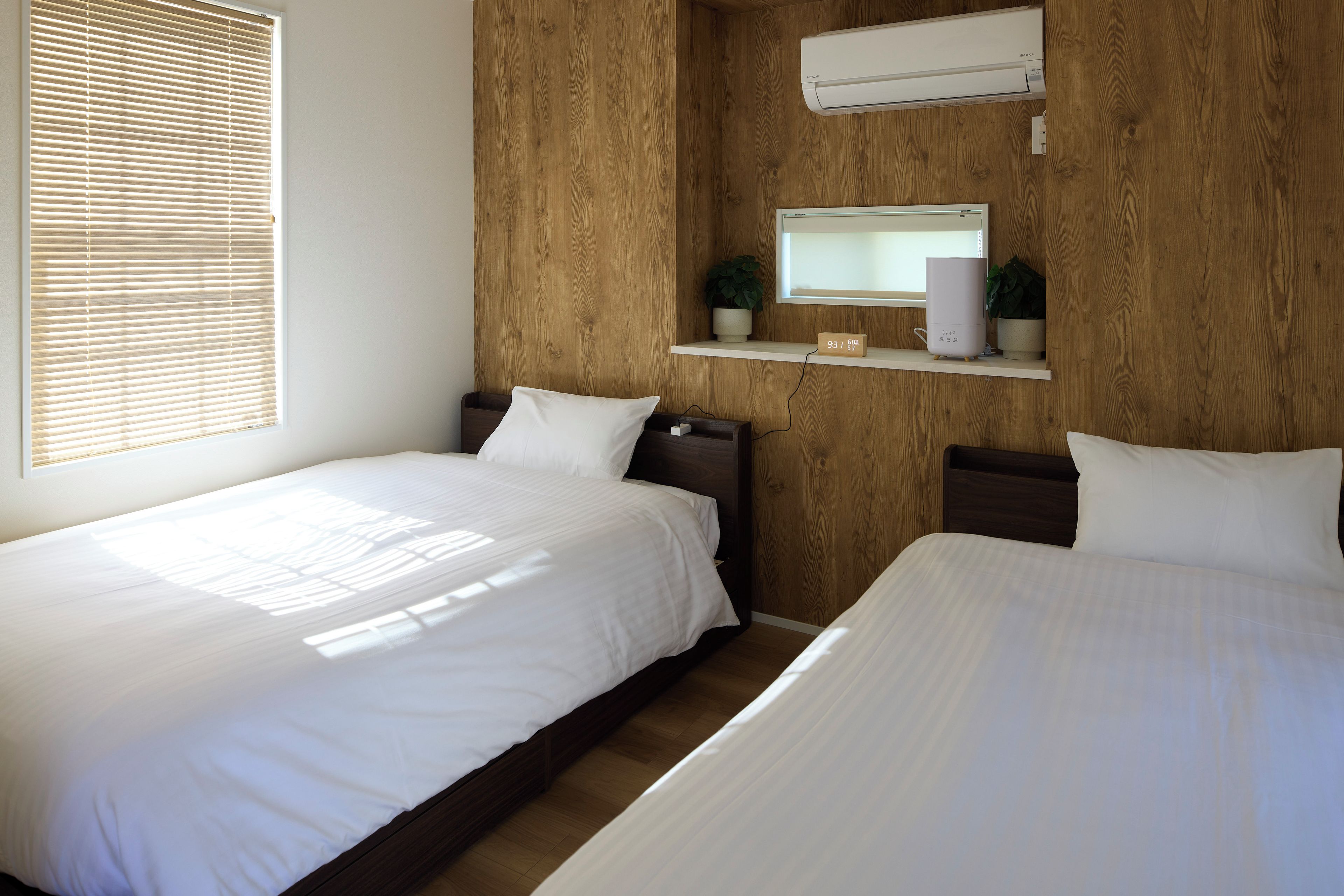 A hotel room with two single beds, a wooden headboard, a wall-mounted air conditioner, a digital clock, and a small shelf with a plant.