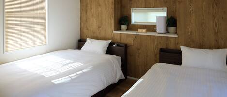 A hotel room with two single beds, a wooden headboard, a wall-mounted air conditioner, a digital clock, and a small shelf with a plant.