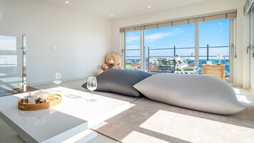 A modern hotel room with a large balcony, a plush sofa, a dining table with wine glasses, and a teddy bear.
