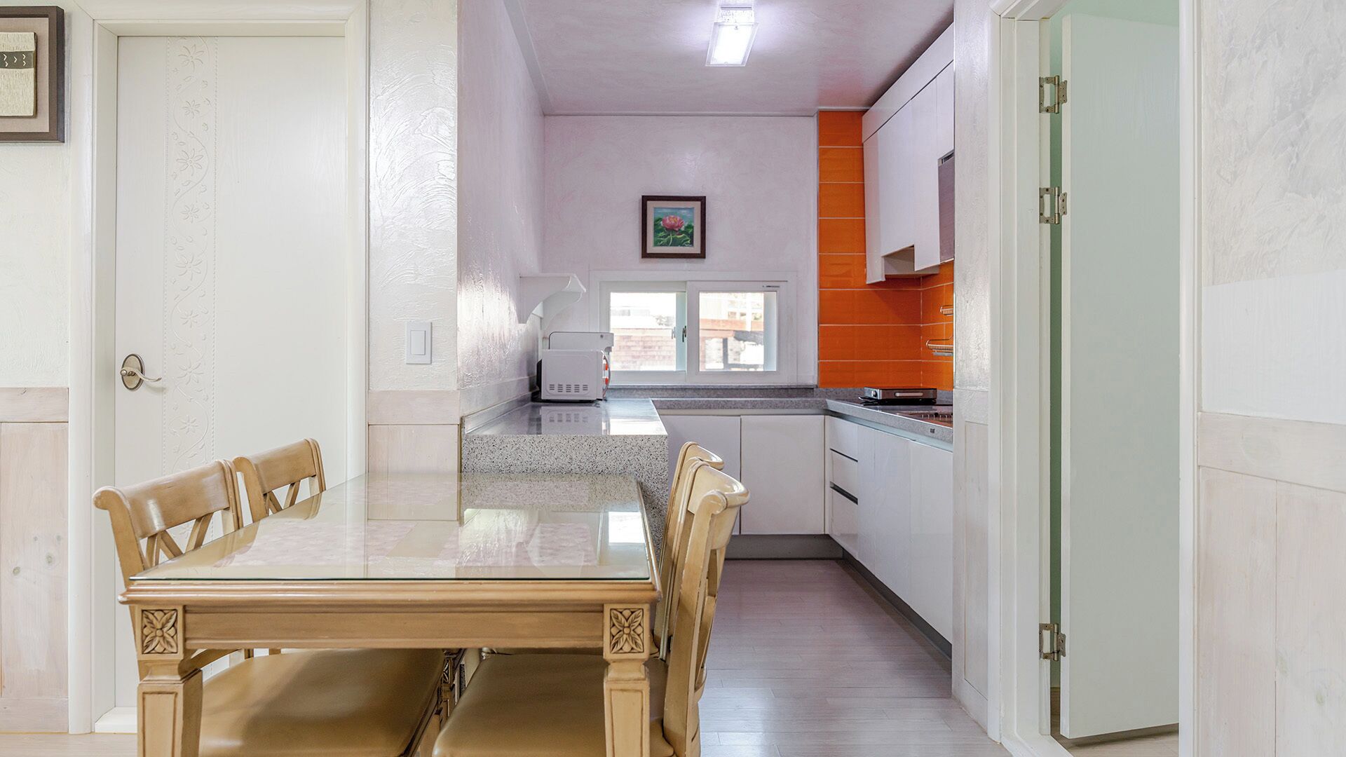 A dining area with a table and chairs, a kitchen with orange cabinets, and a window with a view.