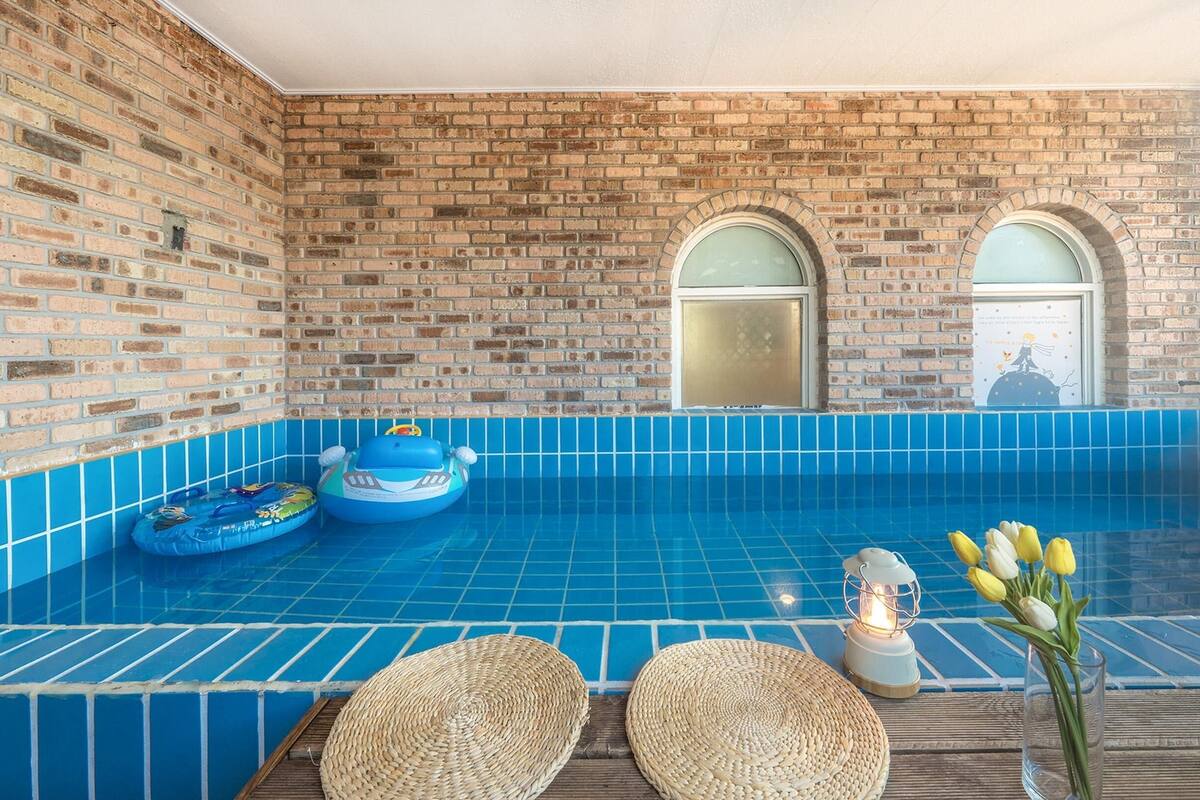 Una piscina con azulejos azules, dos ventanas arqueadas y una terraza de madera con esteras tejidas y un jarrón de flores.