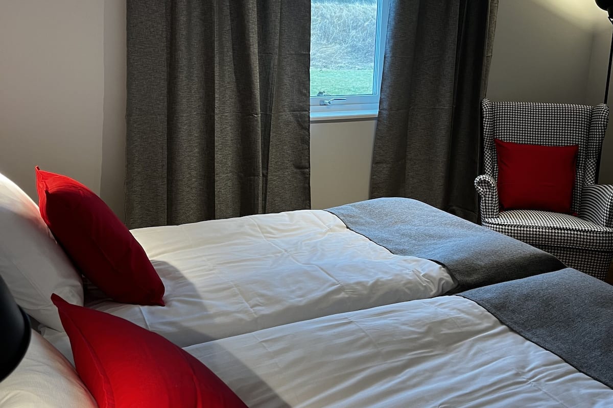 Un dormitorio con dos camas, cortinas grises, una ventana, una silla y una lámpara de pie.