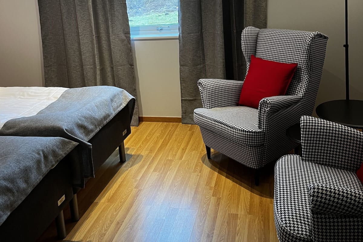 Una habitación con cama, dos sillas, un cojín rojo, una lámpara de pie y una ventana con cortinas.