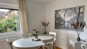 Un comedor con una mesa redonda y sillas blancas, una ventana con vistas a zonas verdes, un cuadro colgado en la pared y un mueble pequeño.