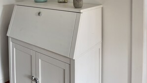 Un mueble blanco con un florero de vidrio que contiene plumas de pampa y una regadera celeste pequeña encima.