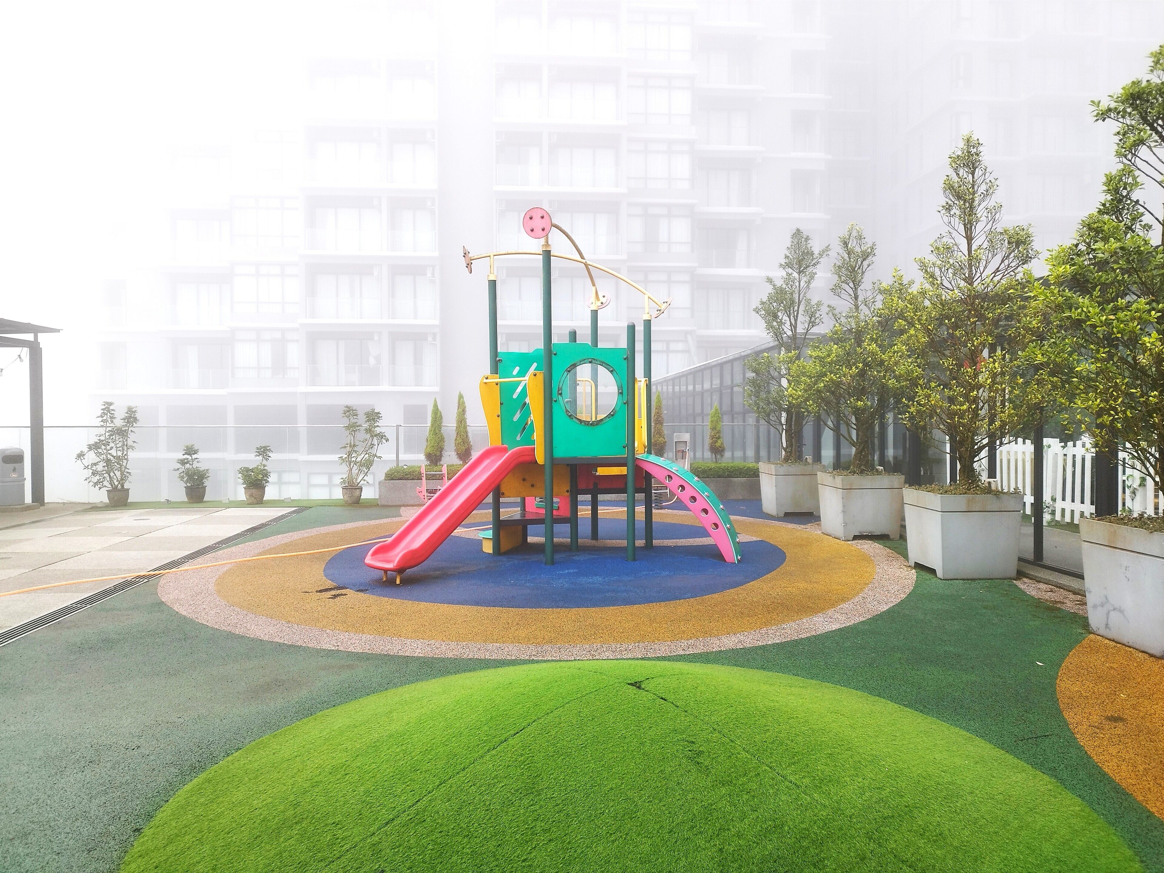 A playground with a slide and climbing structures, surrounded by potted plants and a building in the background.