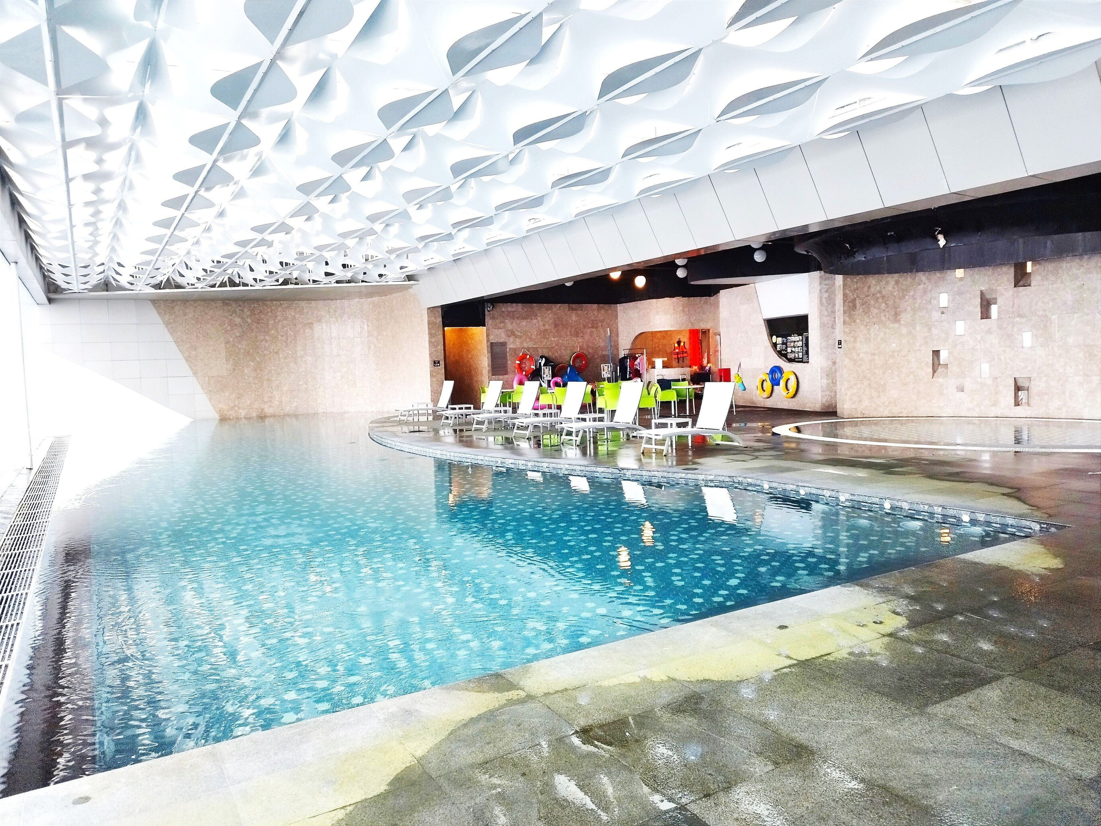An indoor swimming pool with multiple lanes, lounge chairs, and a modern ceiling design.