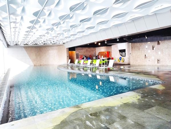 An indoor swimming pool with multiple lanes, lounge chairs, and a modern ceiling design.