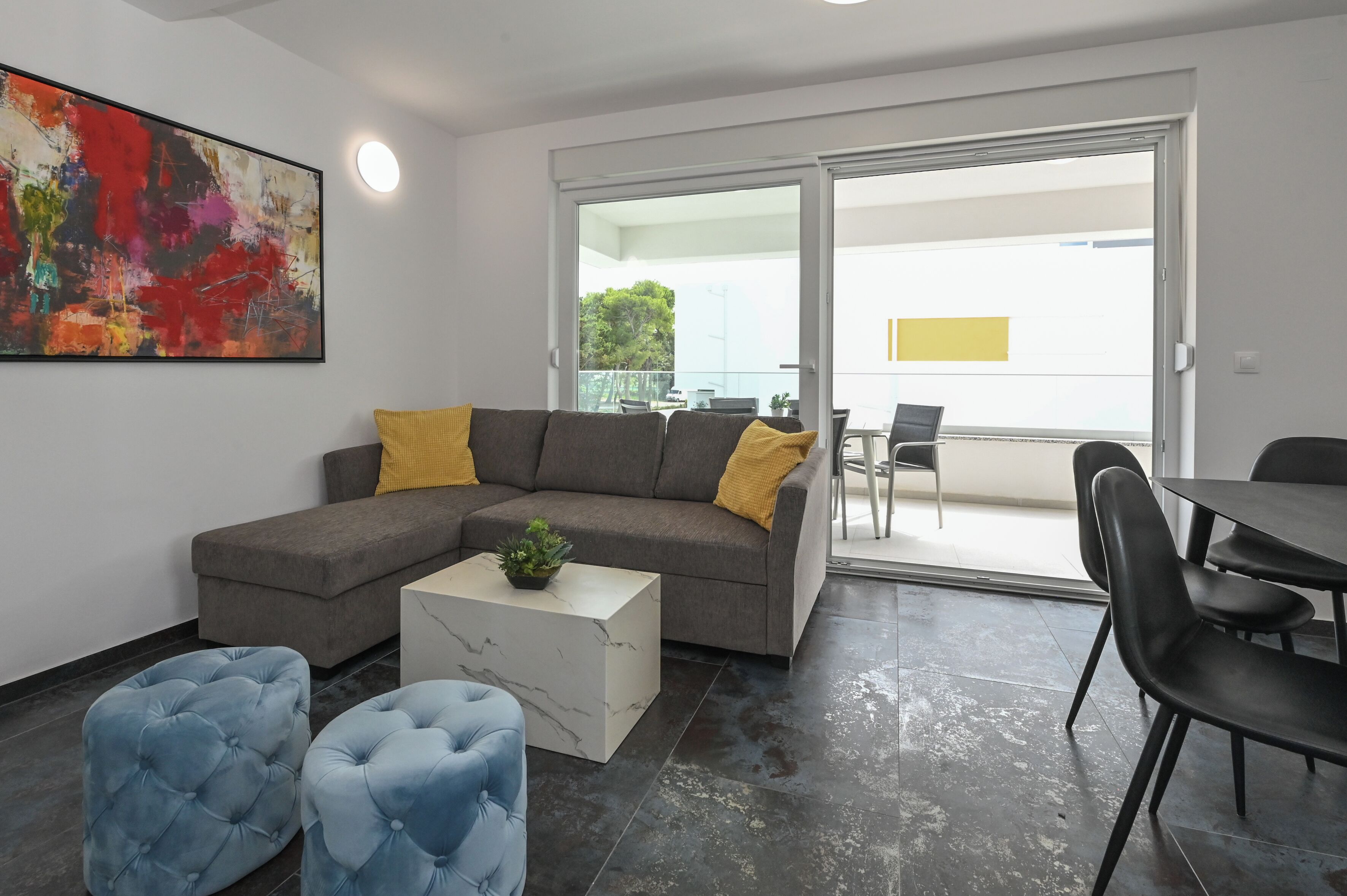 Un salon moderne avec un canapé d’angle gris, des poufs bleus, une table basse en marbre et un tableau abstrait aux couleurs vives.
