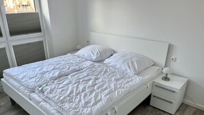 A white bed with a quilted duvet, a bedside table with a lamp, and a drawer.