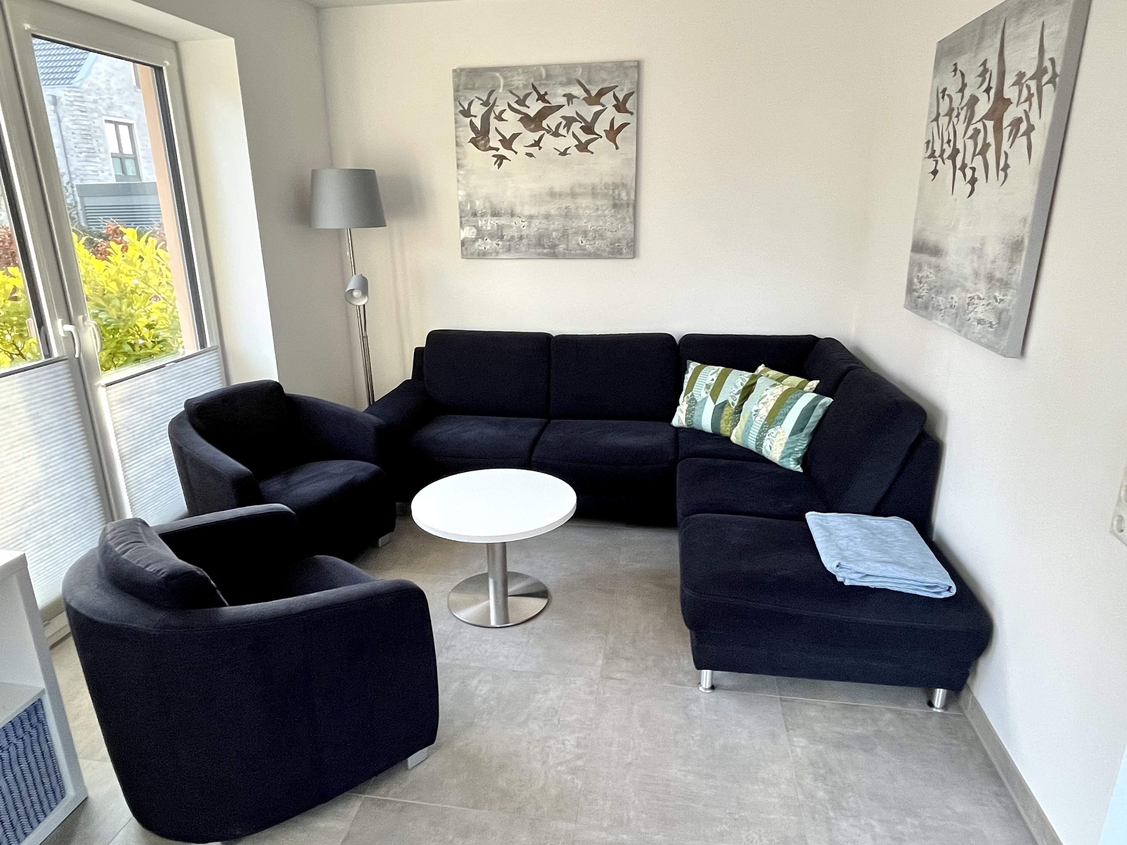 A modern living room with a dark blue sectional sofa, a round coffee table, and two armchairs. There are abstract wall art pieces above the sofa and a floor lamp to the left.