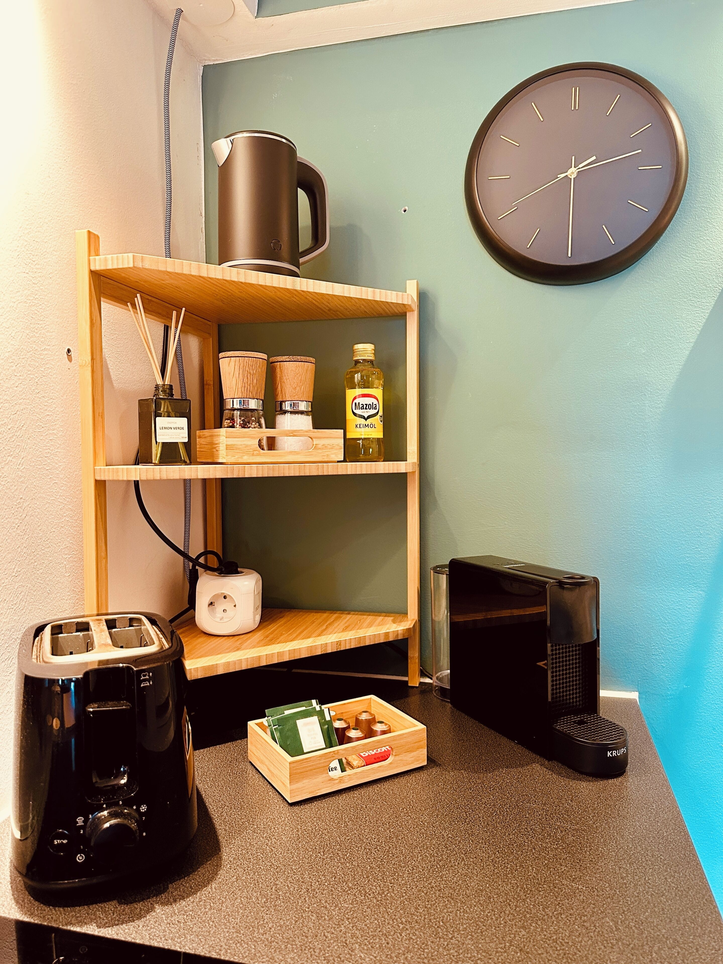 A kitchenette with a toaster, kettle, and coffee maker on the counter, a wooden shelf with kitchen items, and a clock on the wall.