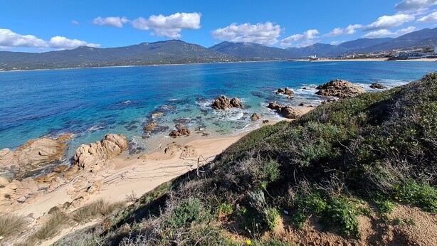 Paisagem costeira com águas azul-claras, litoral rochoso e montanhas ao longe.