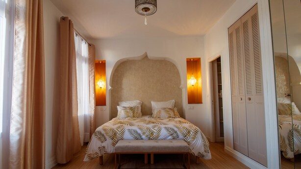 A bedroom with a large bed, a headboard, bedside lamps, a wardrobe, and a hanging light fixture.