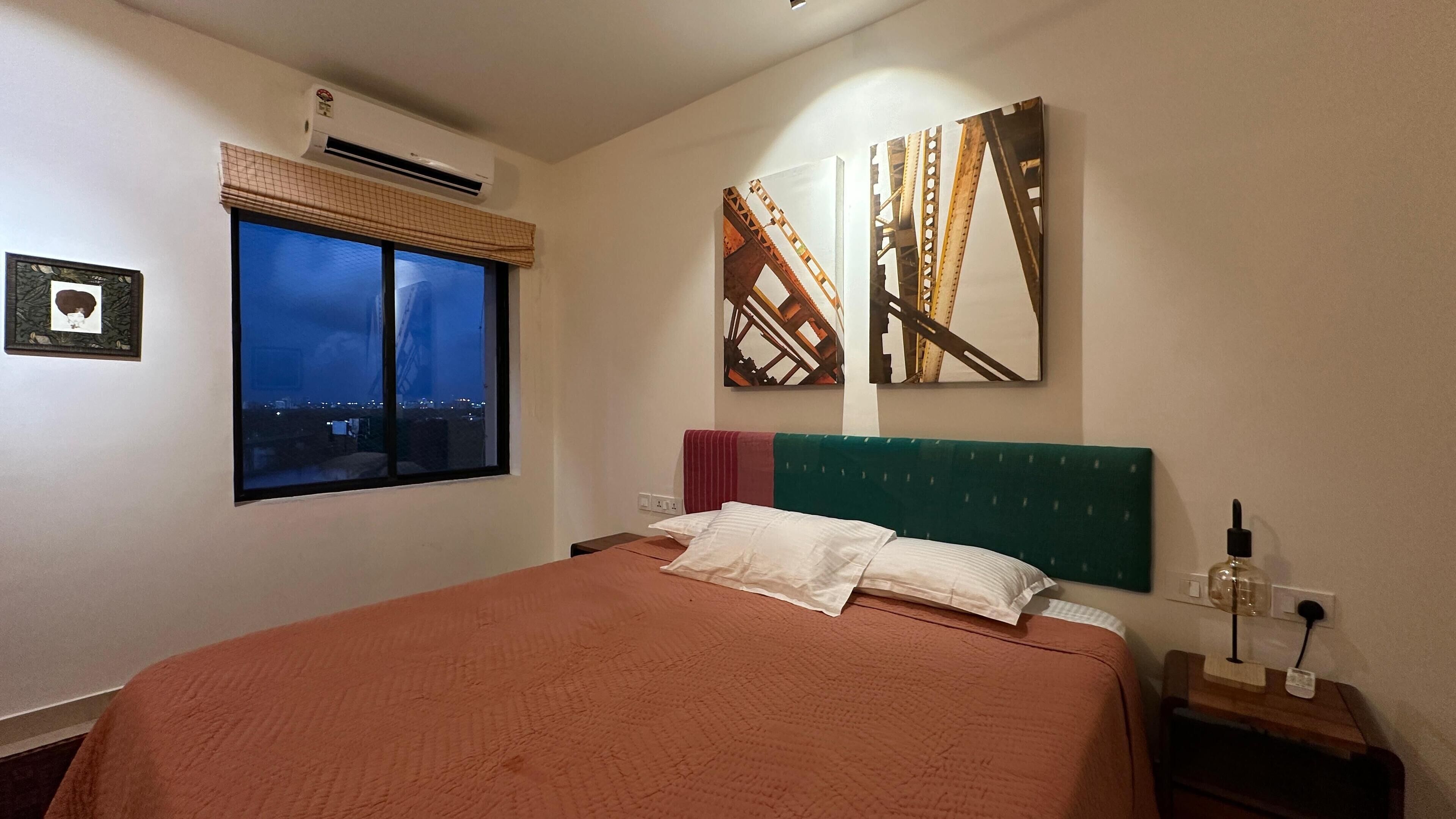 Quarto com cama, janela com vista para a cidade, ar condicionado e obra de arte na parede.