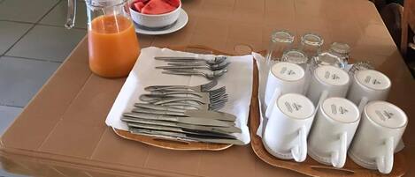 A breakfast table set with utensils, bowls of fruit, a pitcher of orange juice, and several mugs.