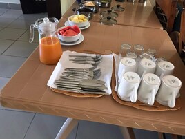 Una mesa de desayuno dispuesta con cubiertos, cuencos de fruta, una jarra de jugo de naranja y varias tazas.