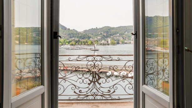 A view from a balcony with a metal railing and glass doors, overlooking a river, buildings, and hills.