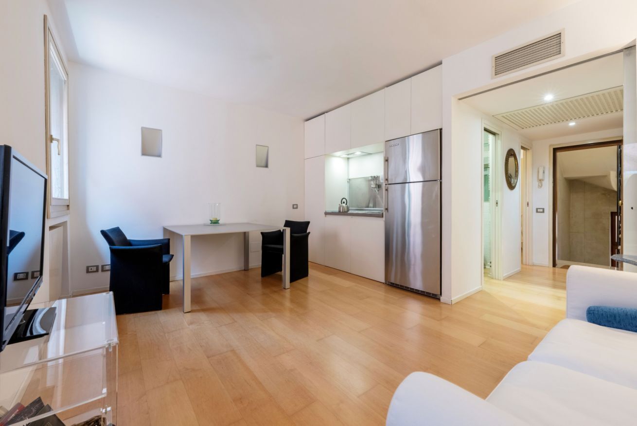 En moderne stue med en hvid sofa, et spiseområde med bord og stole, samt et køkken med køleskab og komfur.