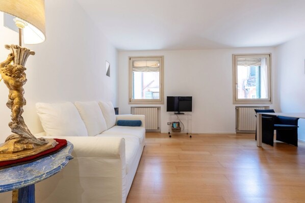 A living room with a white sofa, a small table with a decorative lamp, a TV on a stand, and a dining area with a table and chairs.