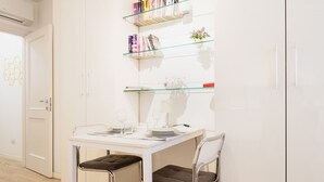 Una habitación pequeña y moderna con una mesa blanca y dos sillas, una estantería con libros y objetos decorativos, y una puerta con un diseño geométrico.