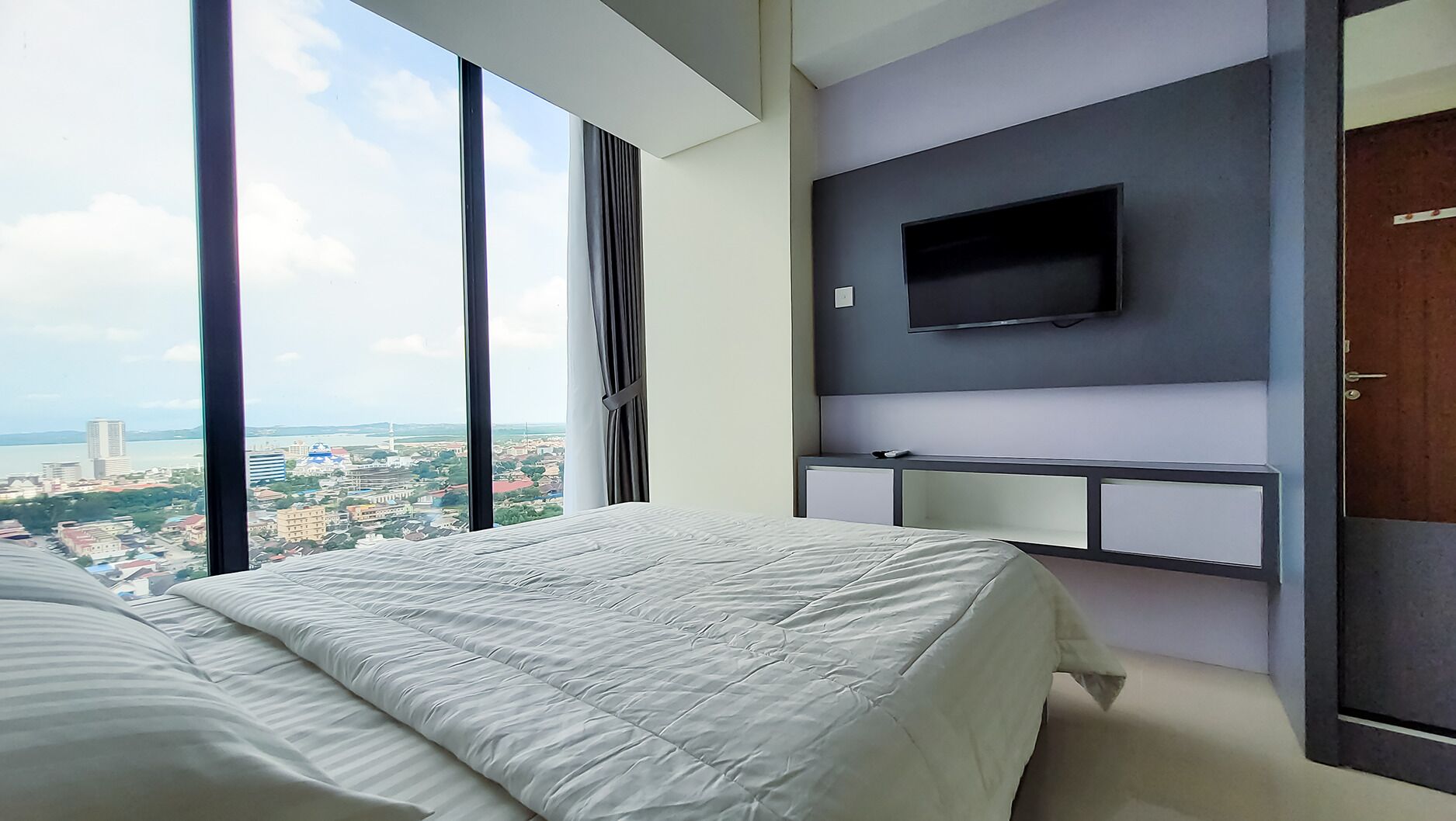 A modern hotel room with a large bed, a TV mounted on the wall, and a view of a cityscape through large windows.