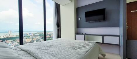 A modern hotel room with a large bed, a TV mounted on the wall, and a view of a cityscape through large windows.