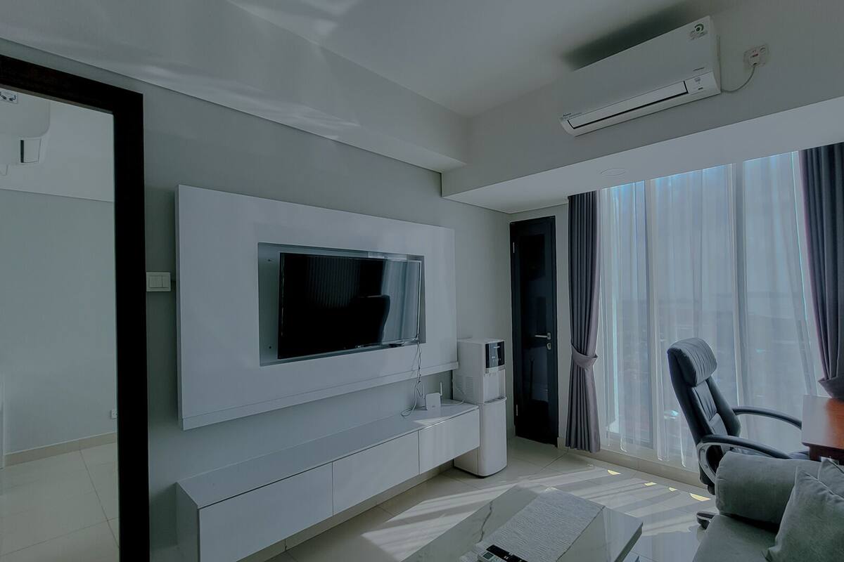 Ein modernes Wohnzimmer mit Wand-Fernseher, einer eleganten weißen Unterhaltungseinheit, einem großen Fenster mit durchsichtigen Vorhängen und einer gemütlichen Sitzecke mit Sofa und Sesseln.
