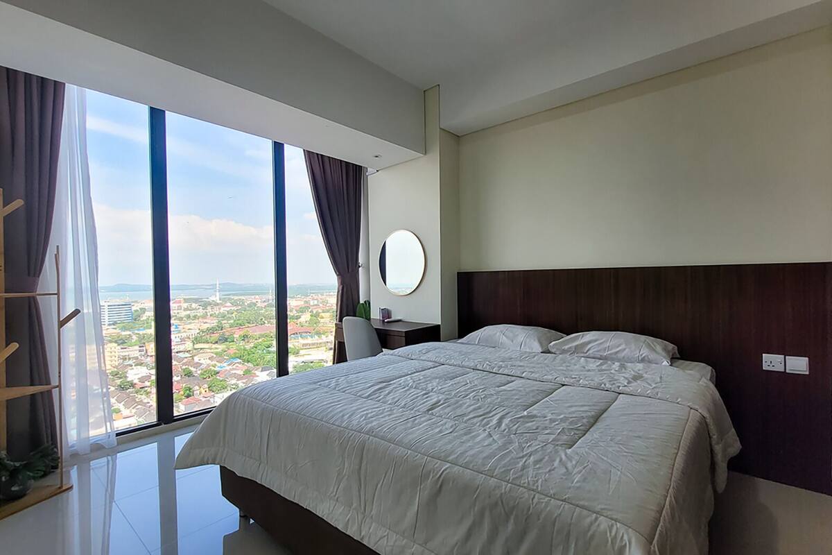 Ein modernes Hotelzimmer mit einem großen Bett, einem Schreibtisch und Blick auf eine Stadtlandschaft durch raumhohe Fenster.