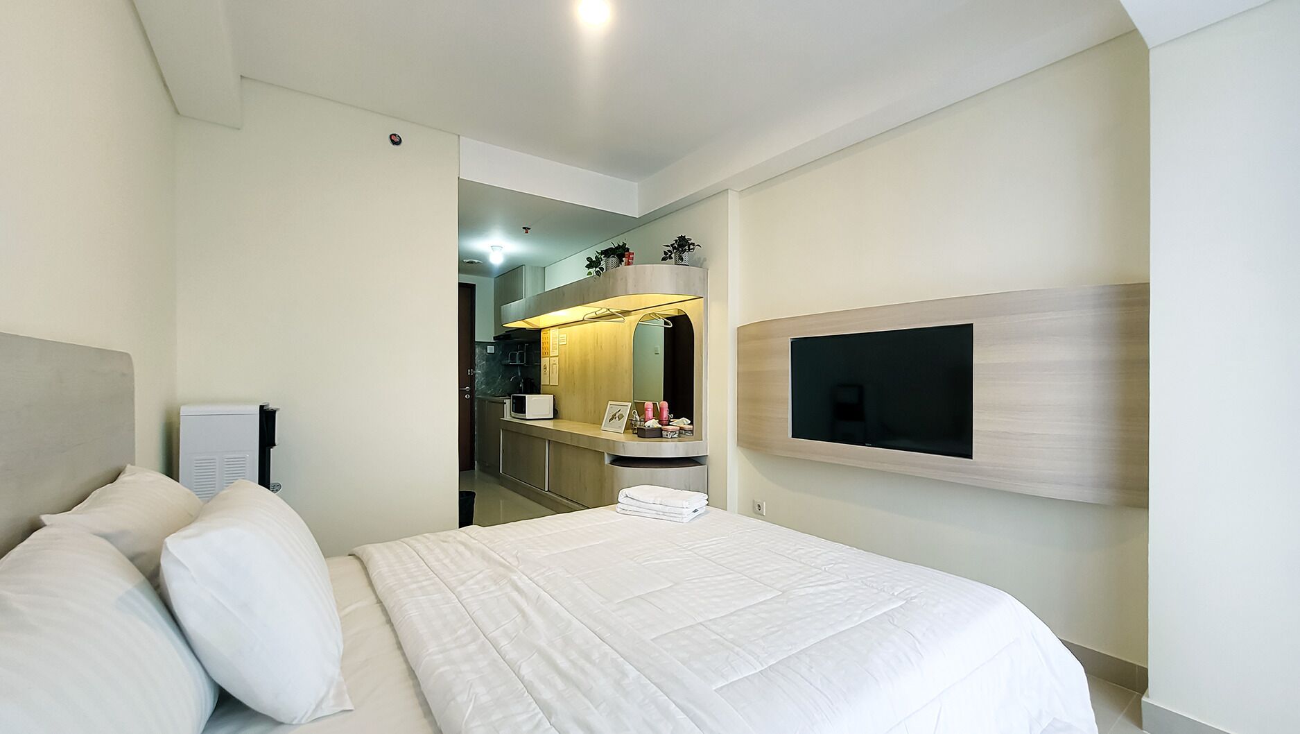 A modern hotel room with a large bed, a flat-screen TV mounted on the wall, and a small kitchenette in the background.