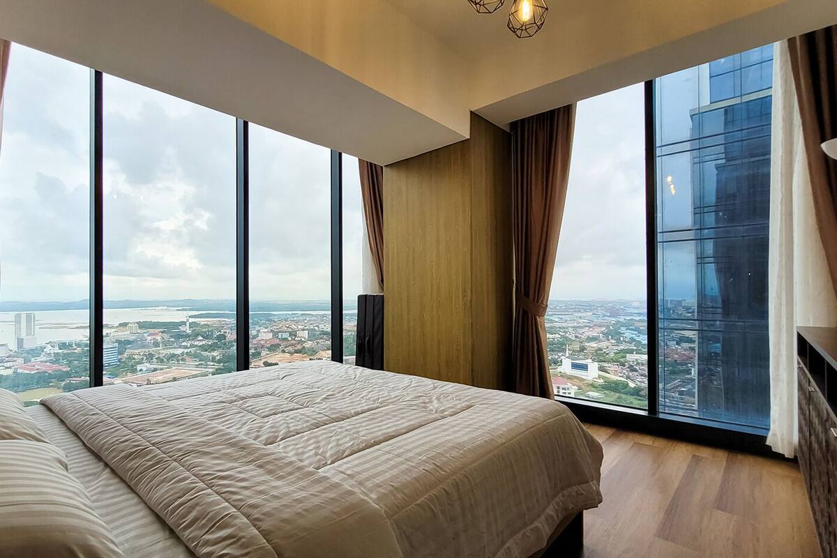 Ein modernes Hotelzimmer mit einem großen Bett, einem Holz-Kopfteil und Blick auf die Stadt durch raumhohe Fenster.