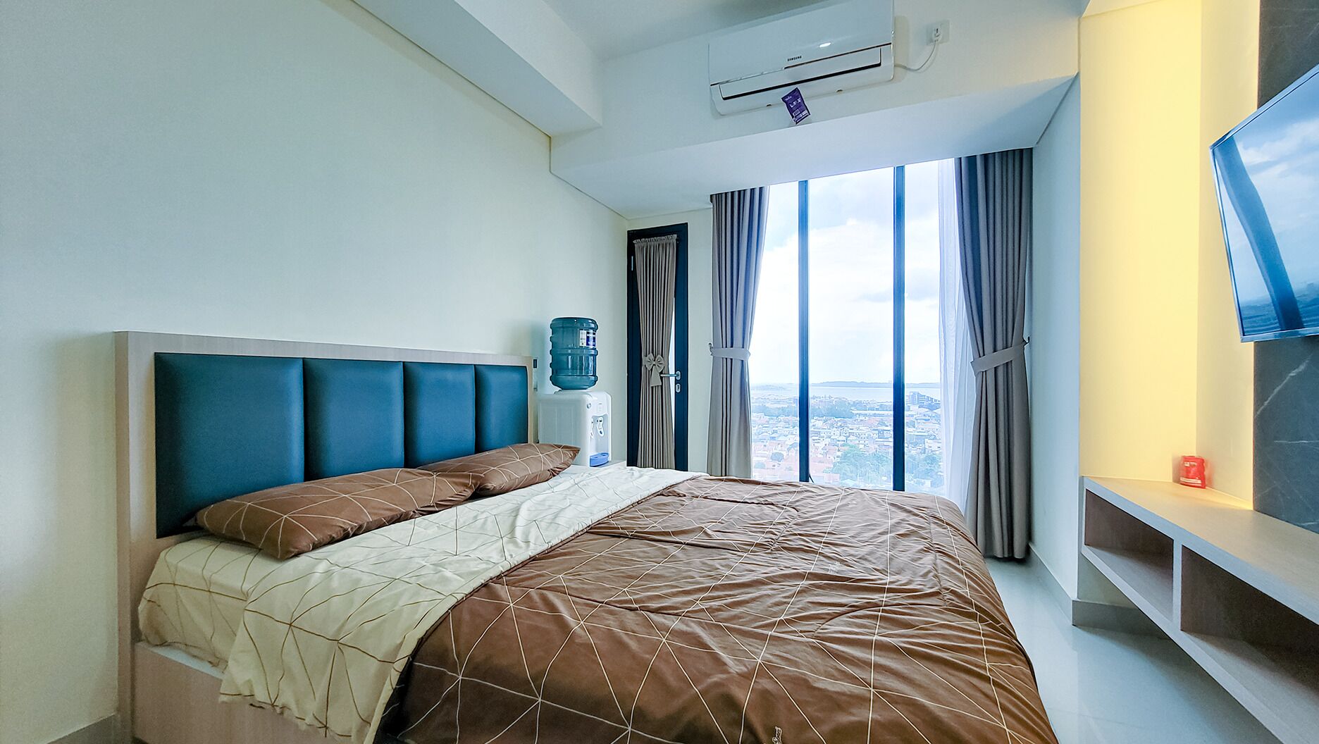 A modern hotel room with a large bed, a water dispenser, a flat-screen TV, and a city view.