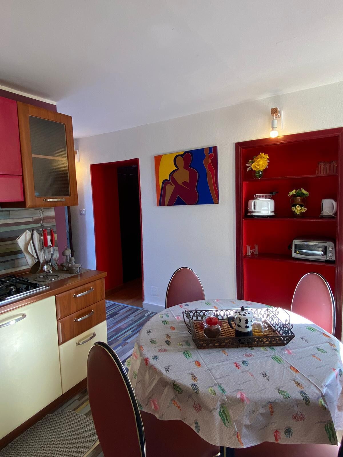 Une cuisine compacte avec un coin repas, comprenant une table recouverte d’une nappe à motifs floraux, des chaises, un meuble rouge avec un grille-pain et un tableau accroché au mur.
