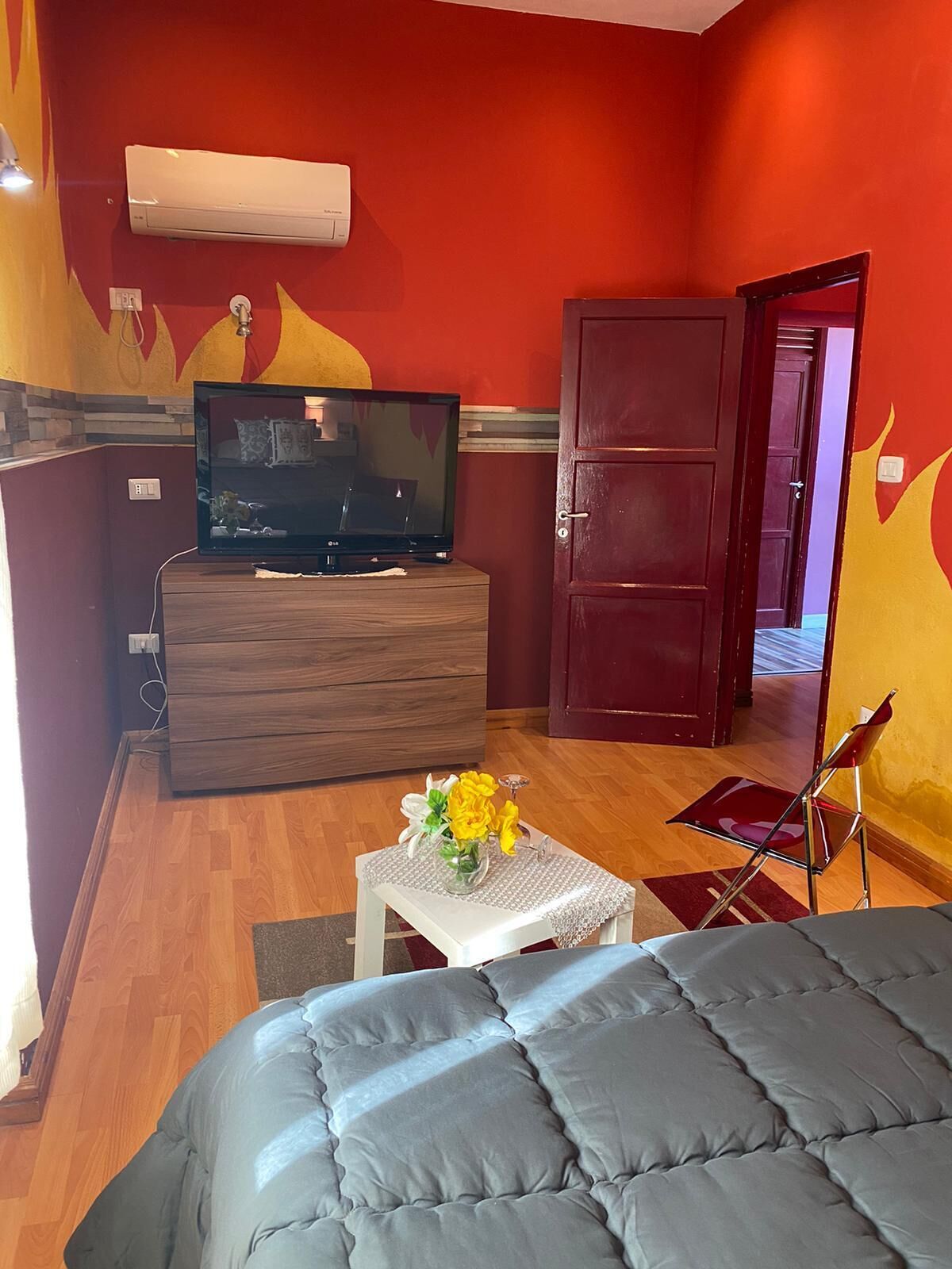 Une pièce avec un lit, un téléviseur posé sur une commode en bois, une petite table avec des fleurs et une porte rouge.