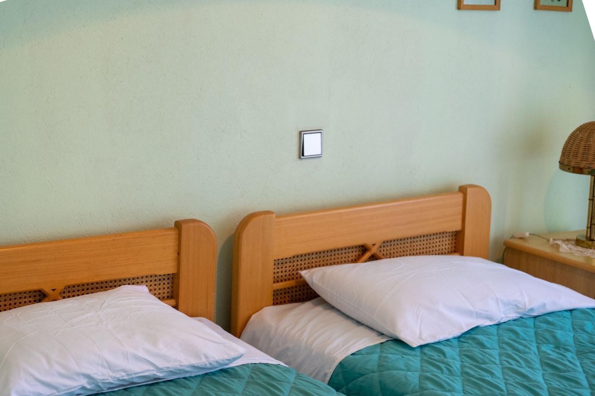 A double bed with wooden headboards and white pillows, a teal bedspread, and a nightstand with a lamp.