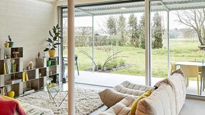Ett modernt vardagsrum med en stor soffa, ett soffbord i glas och en bokhylla. Genom stora fönster har man utsikt över en trädgård.