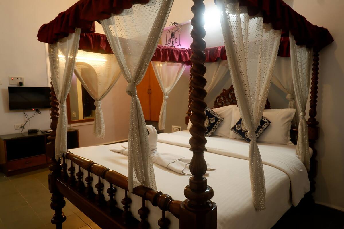 A four-poster bed with white and red curtains, a flat-screen TV on the wall, and a wooden nightstand.