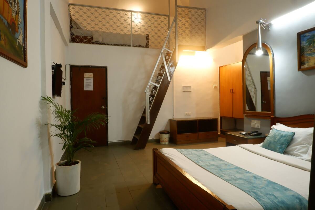 A bedroom with a bed, a bedside table, a nightstand, a mirror, a potted plant, a door, and a staircase.