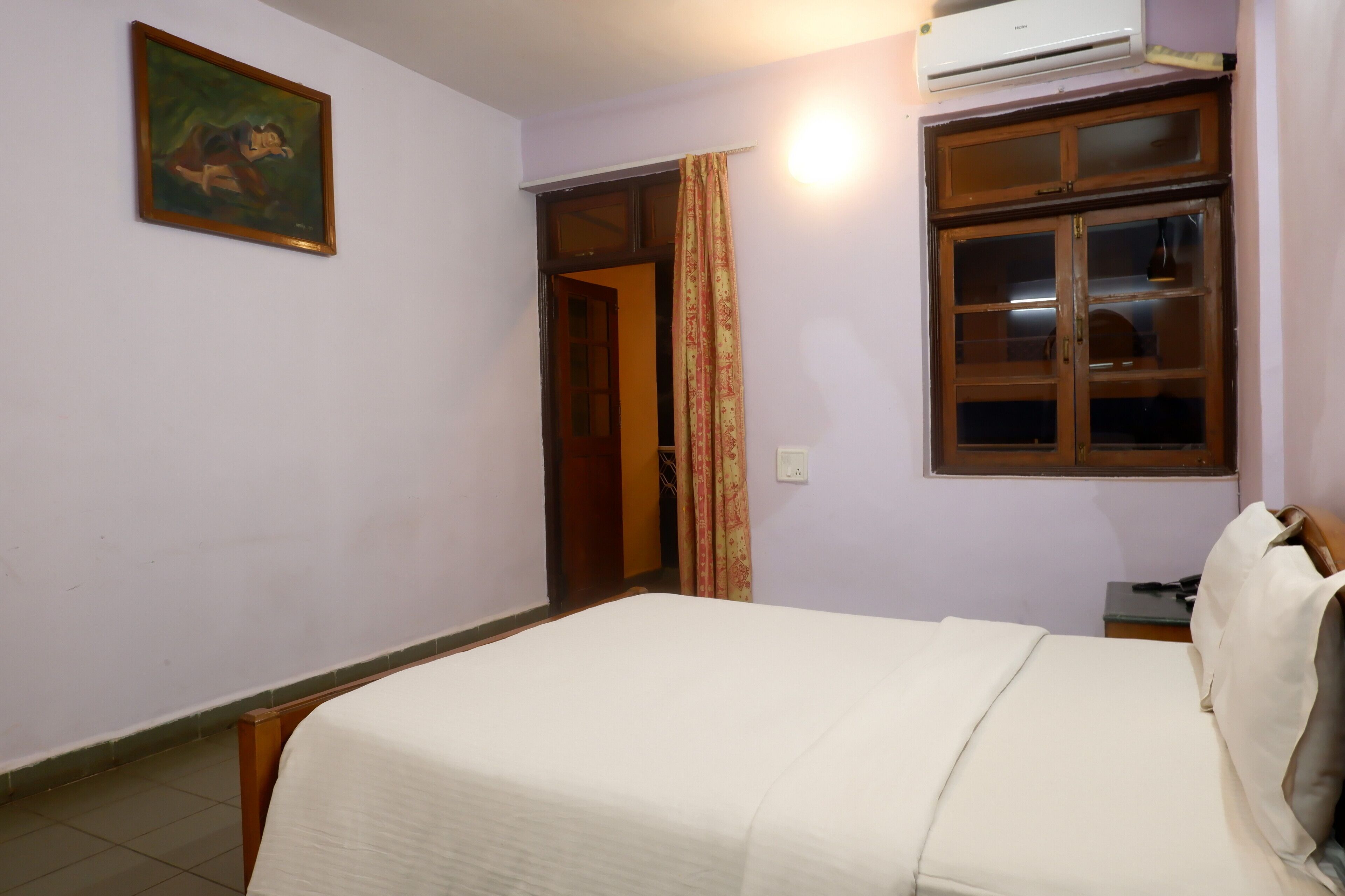 A bedroom with a bed, an air conditioning unit, a window with curtains, and a door leading to another room.