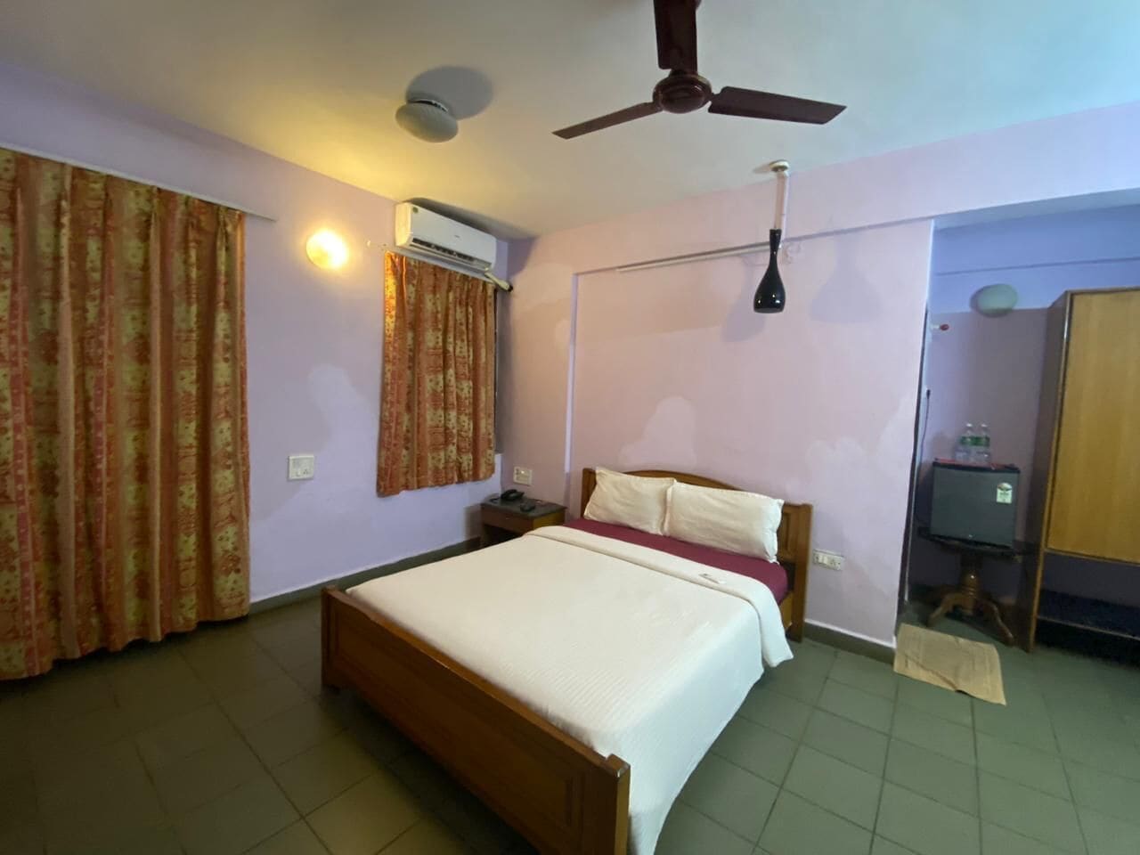 A hotel room with a single bed, a ceiling fan, an air conditioning unit, a bedside table, and a wardrobe.