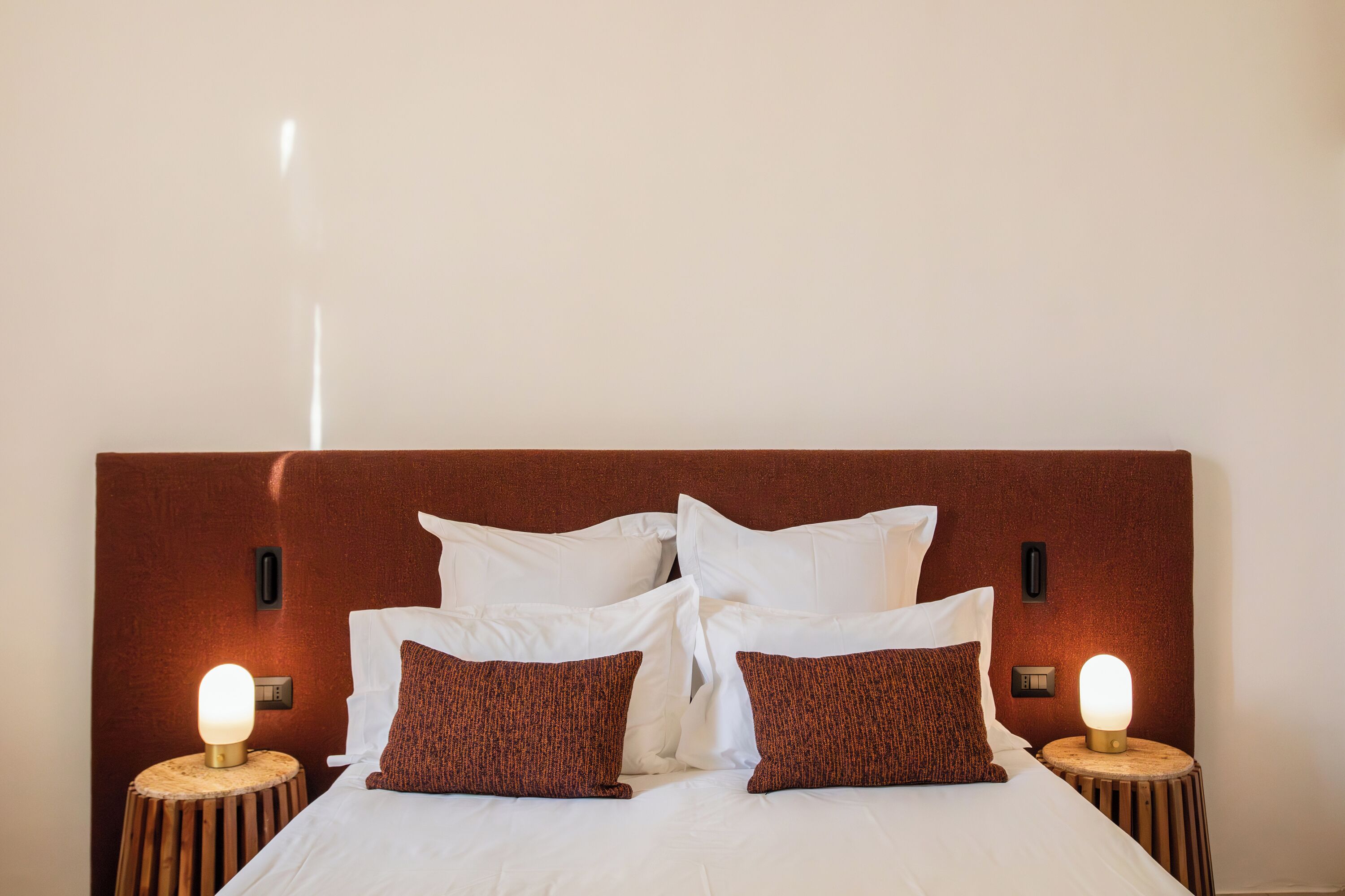 Una cama bien tendida con sábanas blancas y dos almohadas marrones, con dos mesitas de noche de madera que tienen lámparas iluminadas.